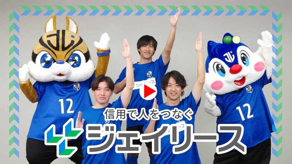 ジェイリースFCのCMのサムネイル画像。中央にジェイリースFCの選手が3人、その両側にはジェイリースFCのマスコットキャラクターが1体づつ立っており、キャラクターと選手はアルファベットのJを表すように、左手を頭上へ高く上げ、右手は肩の高さにあげるポーズをとっている。画面の下部には「信用で人をつなぐジェイリース」というテキストがあり、動画の再生ボタンを表すアイコンが画像の中央に置かれている。動画は新しいウィンドウで開く。